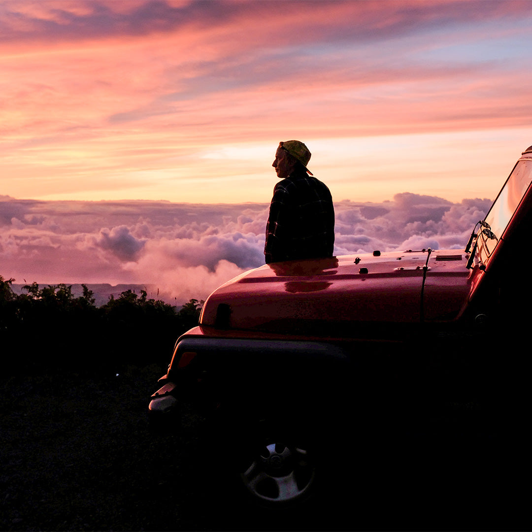 A person sits on the hood of a red SUV, gazing at a vibrant sunset above clouds and distant hills. The scene exudes tranquility and adventure.