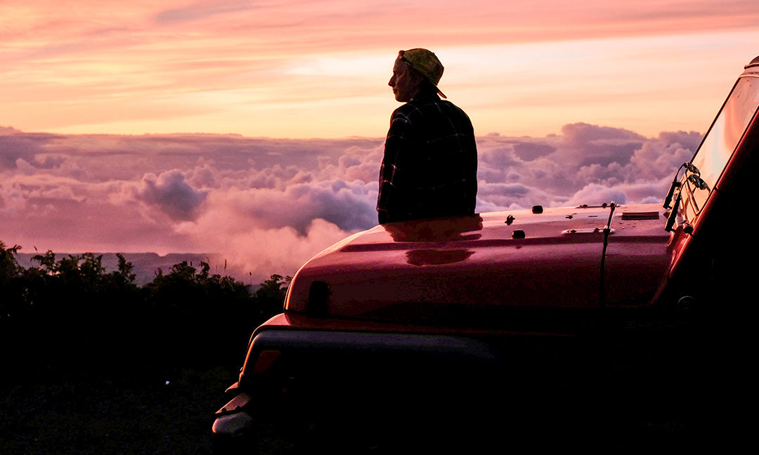 A person sits on the hood of a red SUV, gazing at a vibrant sunset above clouds and distant hills. The scene exudes tranquility and adventure.