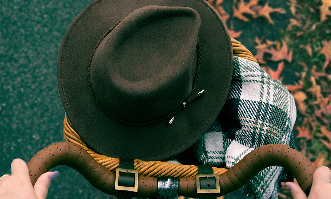 A person holds the handlebars of a bicycle with a brown hat and a plaid scarf in the front basket. Fallen autumn leaves are scattered on the ground ahead.