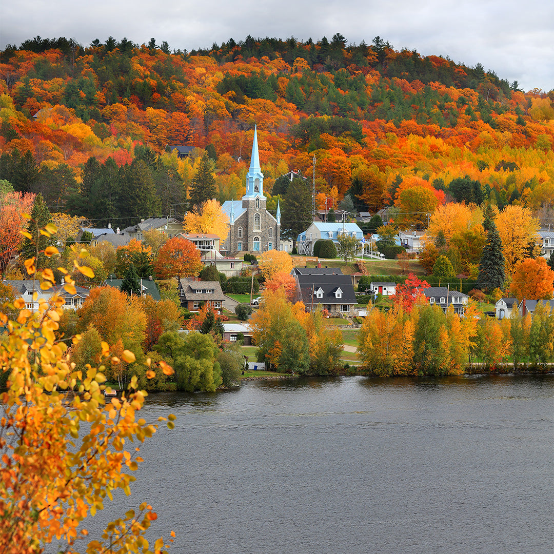 A quaint village with a prominent church steeple sits by a river, surrounded by vibrant autumn trees in shades of orange, red, and yellow on rolling hills.