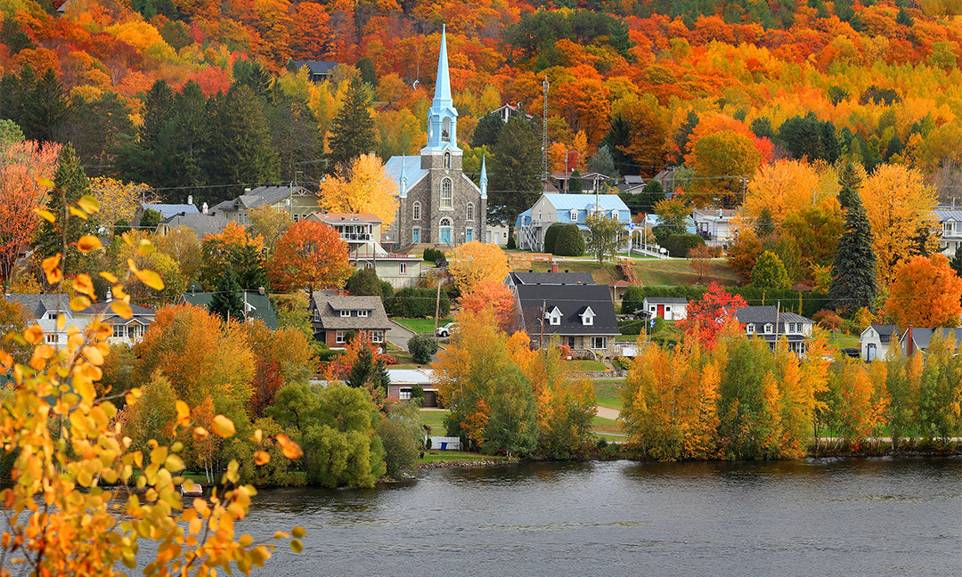 A quaint village with a prominent church steeple sits by a river, surrounded by vibrant autumn trees in shades of orange, red, and yellow on rolling hills.