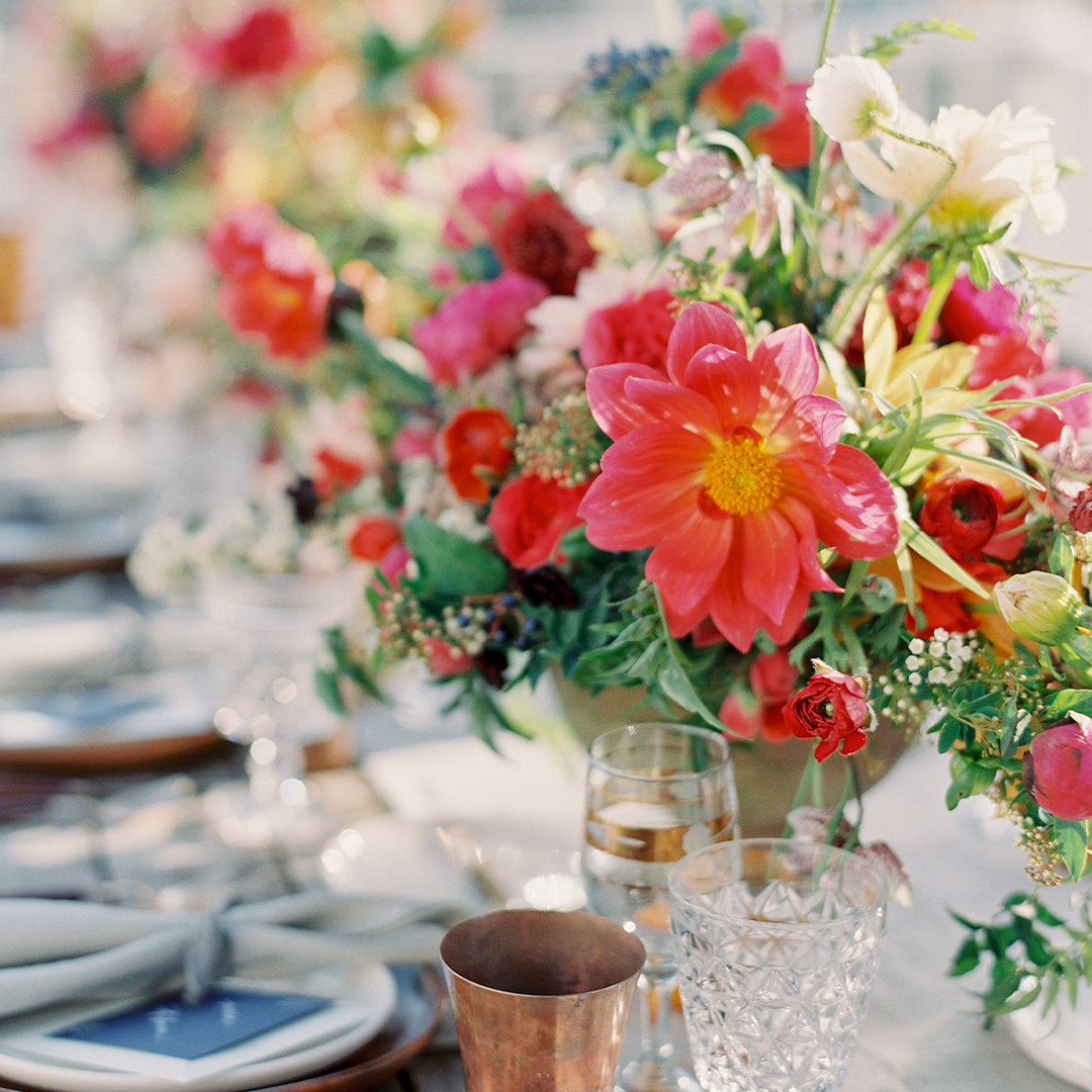 A vibrant table centerpiece with pink, red, and yellow flowers, surrounded by glassware, a copper cup, and neatly arranged plates with napkins, creating an elegant and festive setting.