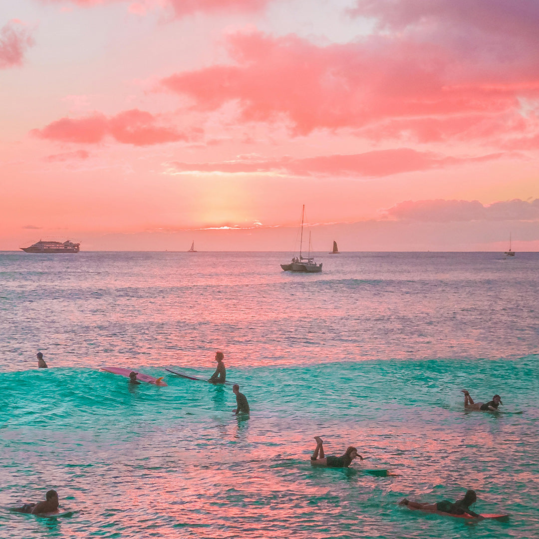 People swimming and surfing in turquoise waves at sunset, with sailboats and a distant cruise ship on the horizon under a pink and orange sky.