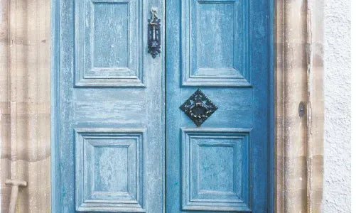 A pair of tall, ornate blue wooden doors with decorative black metalwork at the bottom and a metal knocker, set in a stone doorway.