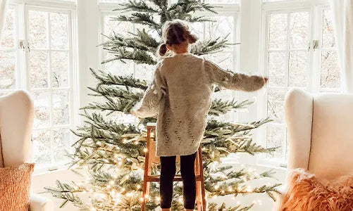 A young child stands barefoot on a wooden step stool, decorating a Christmas tree in a bright, cozy living room with large windows, white curtains, and two armchairs with pillows.