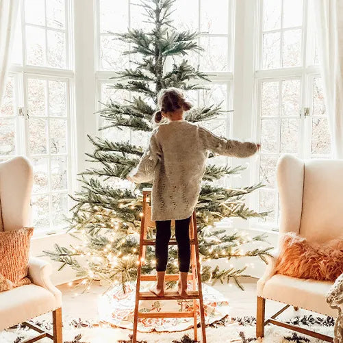 A young child stands barefoot on a wooden step stool, decorating a Christmas tree in a bright, cozy living room with large windows, white curtains, and two armchairs with pillows.
