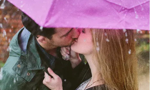 A couple kisses under a pink umbrella in the rain, shielding themselves from the falling drops. The background is softly blurred, highlighting their romantic moment.