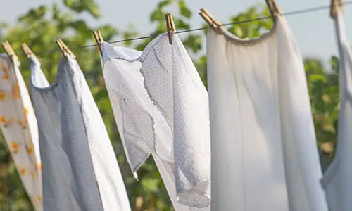 Freshly washed white and light-colored clothes hanging on a clothesline with wooden clothespins, drying outdoors on a sunny day with greenery in the background.