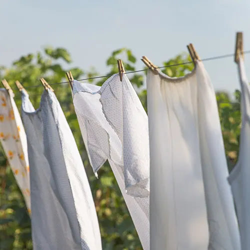 Freshly washed white and light-colored clothes hanging on a clothesline with wooden clothespins, drying outdoors on a sunny day with greenery in the background.