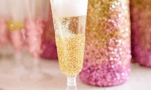 A champagne flute coated with gold and pink glitter sits on a white surface, filled with a bubbly drink and topped with foam. Blurred glittery bottles and glasses appear in the background.