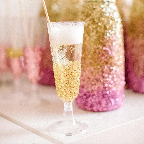 A champagne flute coated with gold and pink glitter sits on a white surface, filled with a bubbly drink and topped with foam. Blurred glittery bottles and glasses appear in the background.