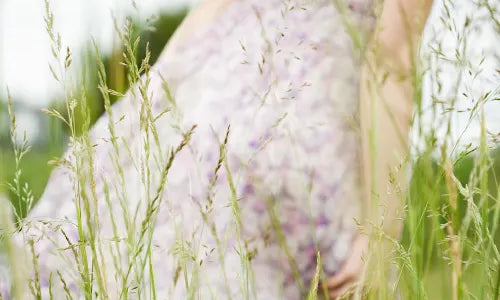 A person in a flowy, light purple floral dress stands in tall grass, with their back turned and hand holding the dress, in a blurred, dreamy outdoor setting.