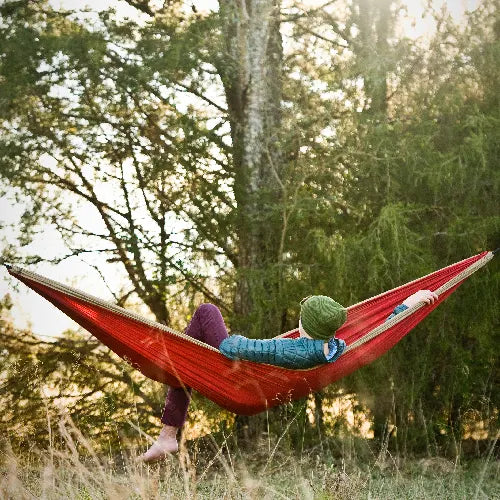 A person wearing a green hat relaxes in a red hammock outdoors, surrounded by tall trees and soft sunlight filtering through the branches.