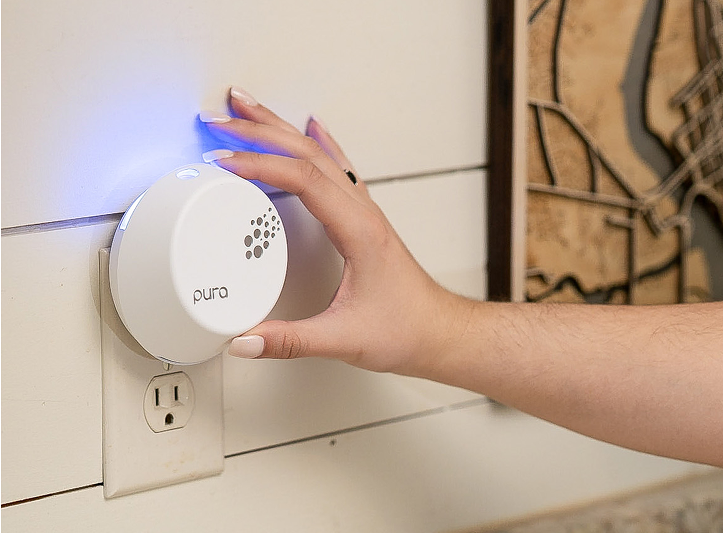 A persons hand adjusts a white Pura air freshener plugged into a wall outlet, with a blue indicator light glowing. The wall is white with horizontal panels, and part of a decorative map is visible.