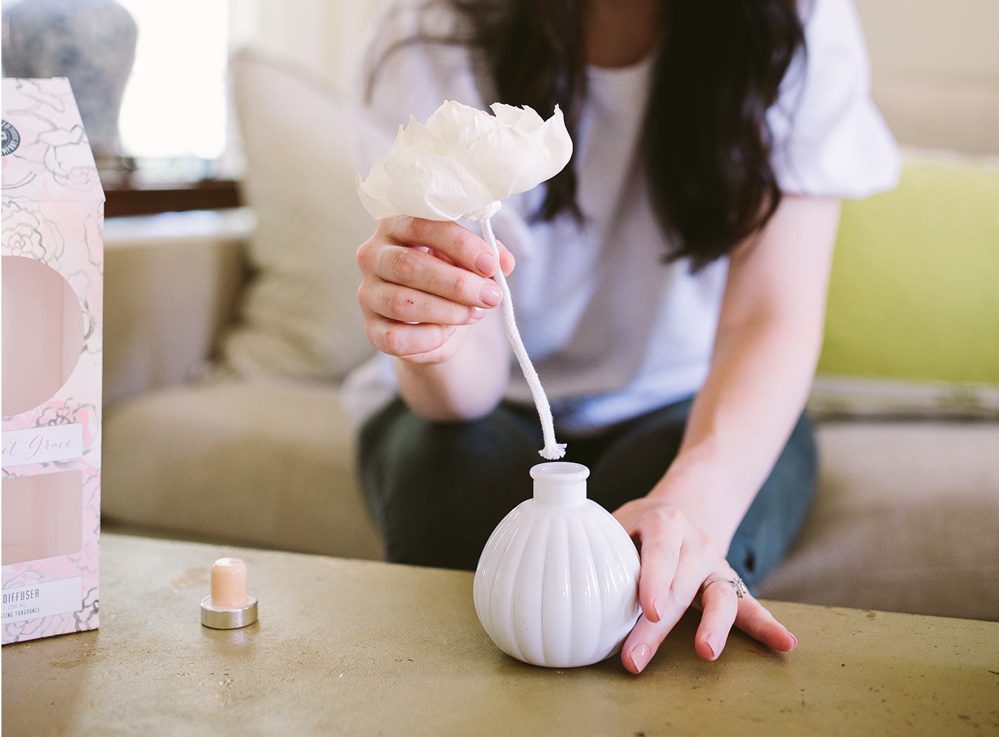 A person places a white paper flower with a stem into a small, round, white vase on a table. A small candle and a decorative box sit nearby, with a sofa and cushions in the background.