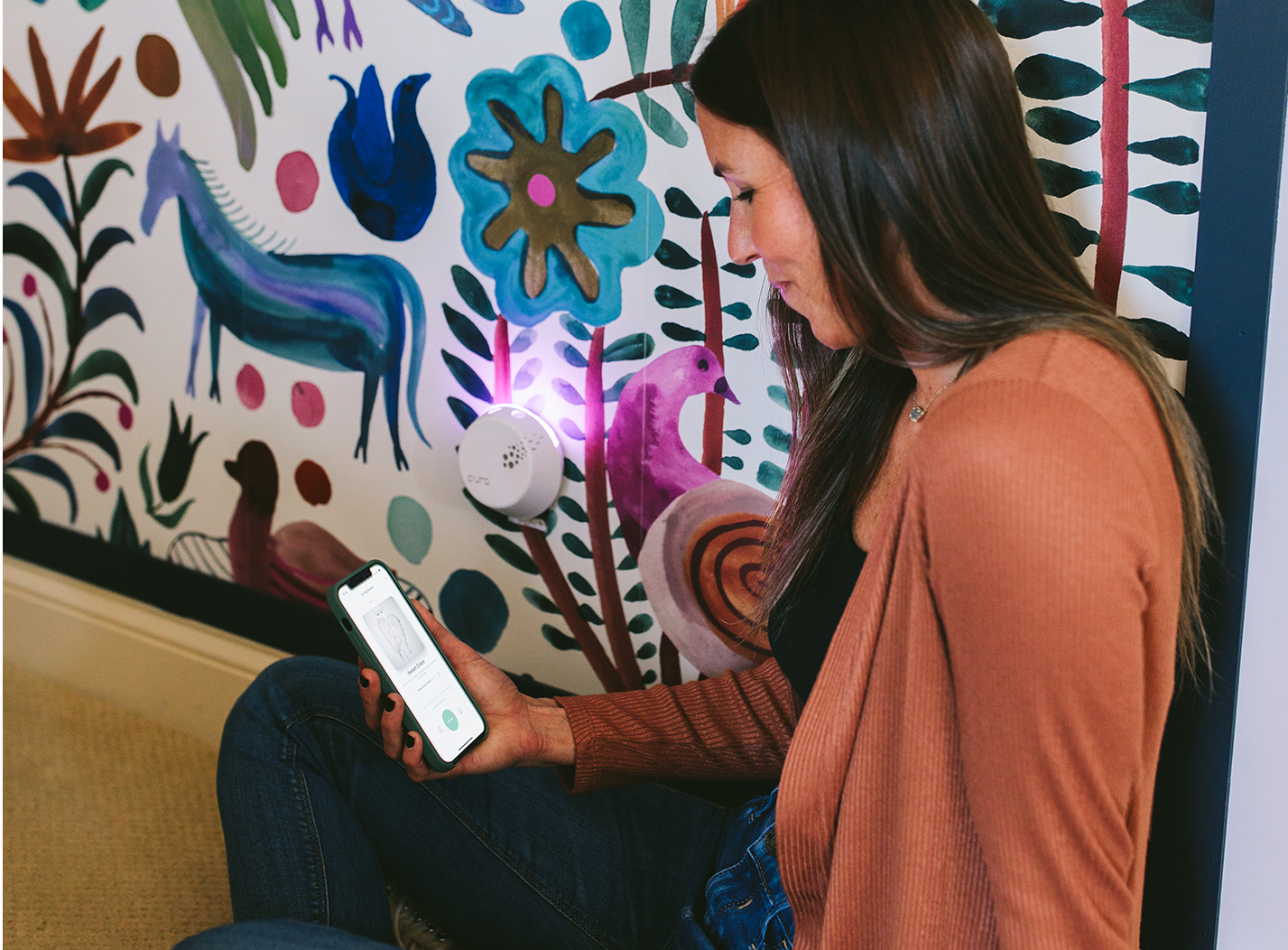 A woman in a brown sweater sits on the floor by a colorful, animal-themed mural, looking at her smartphone. A round electronic device with a glowing light is plugged into the wall next to her.