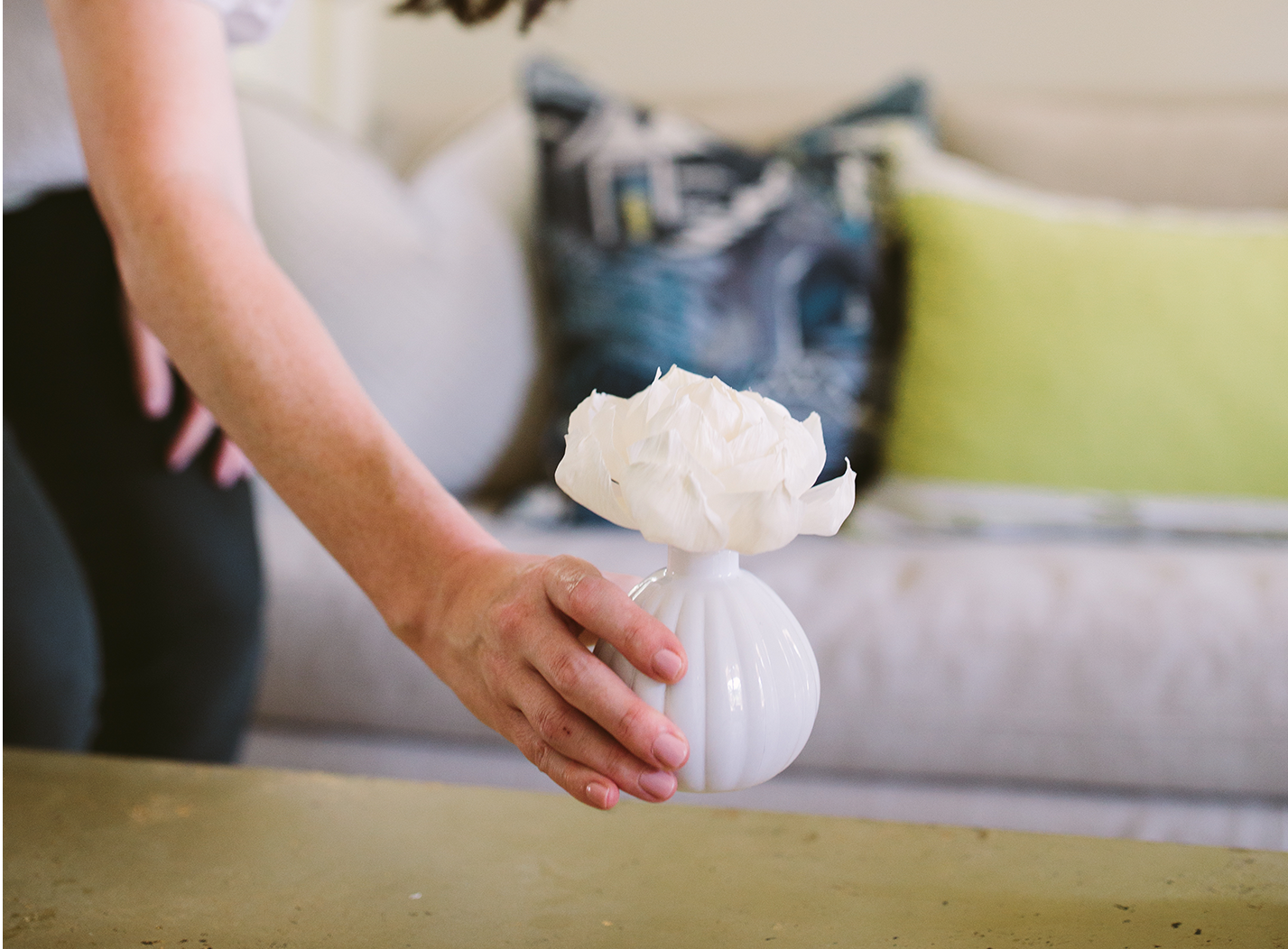 A person places a small white vase with a single white flower on a table. In the background, there is a white couch with blue and green pillows.