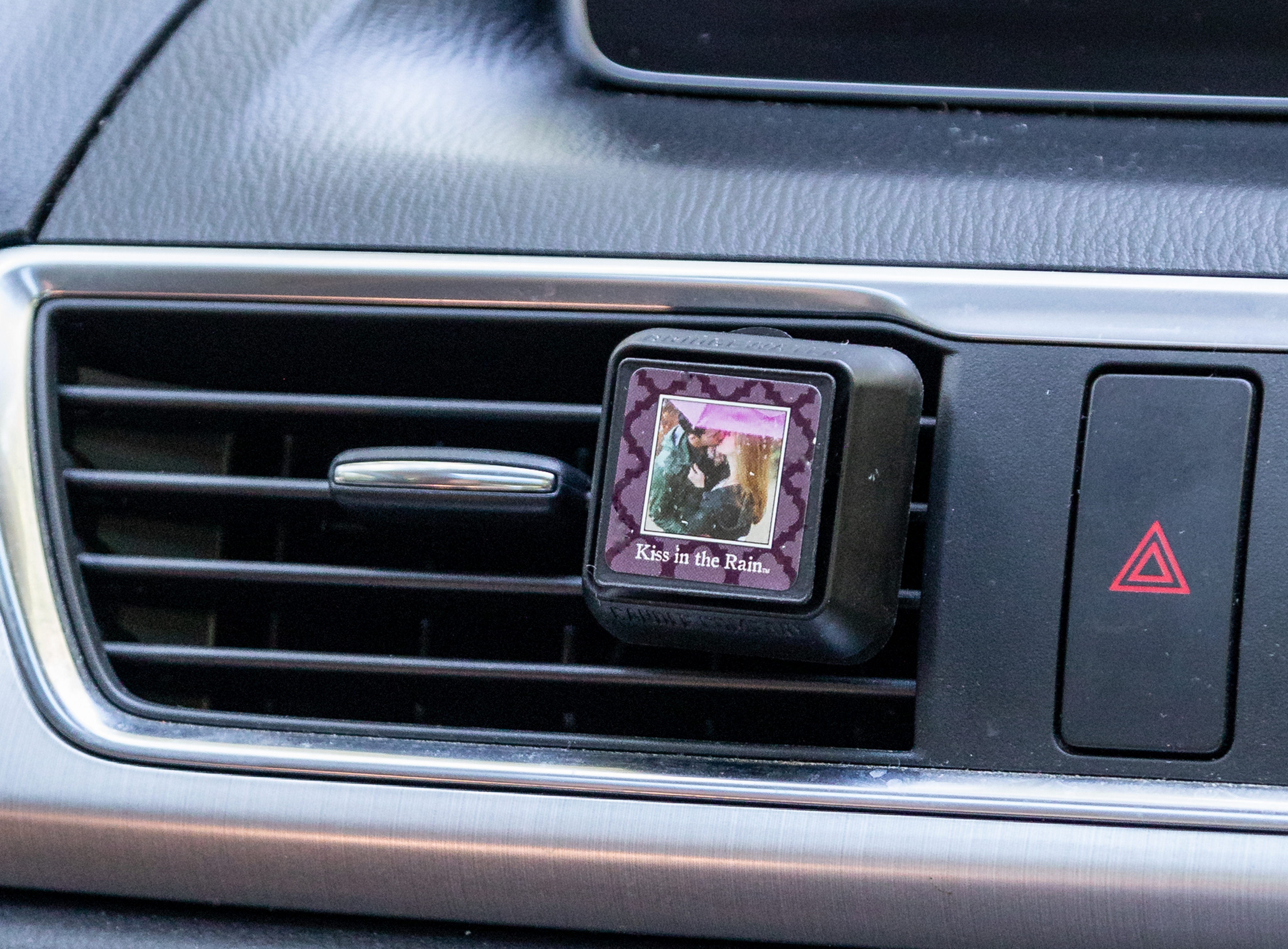 A close-up of a car’s air vent with a square air freshener attached. The freshener has an image of a couple kissing and the words “Kiss in the Rain” printed below the image.