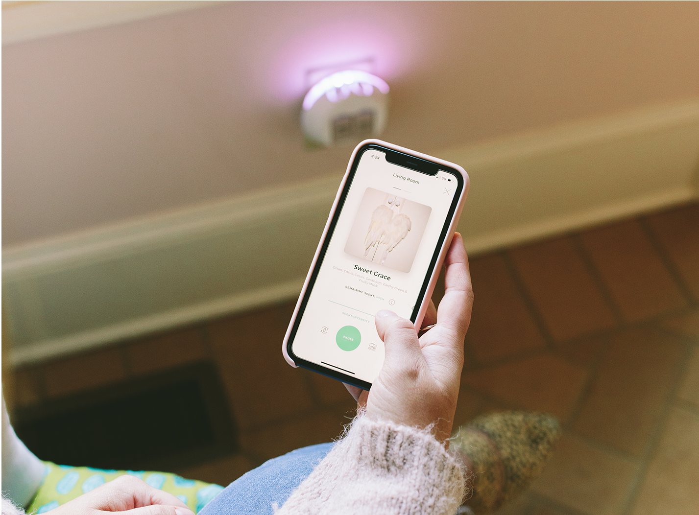 A person wearing a light pink sweater holds a smartphone displaying a music app with a song called Sweet Cora and an image of angel wings. A plugged-in nightlight glows on the wall in the background.
