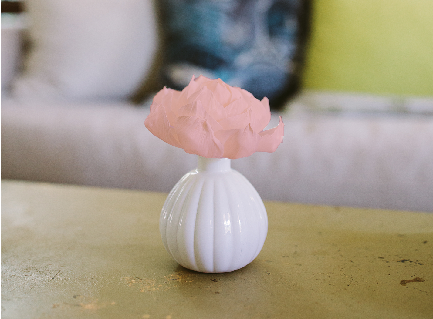 A small white vase with vertical ridges holds a single light pink flower, placed on a beige surface with a blurred couch and colorful cushions in the background.