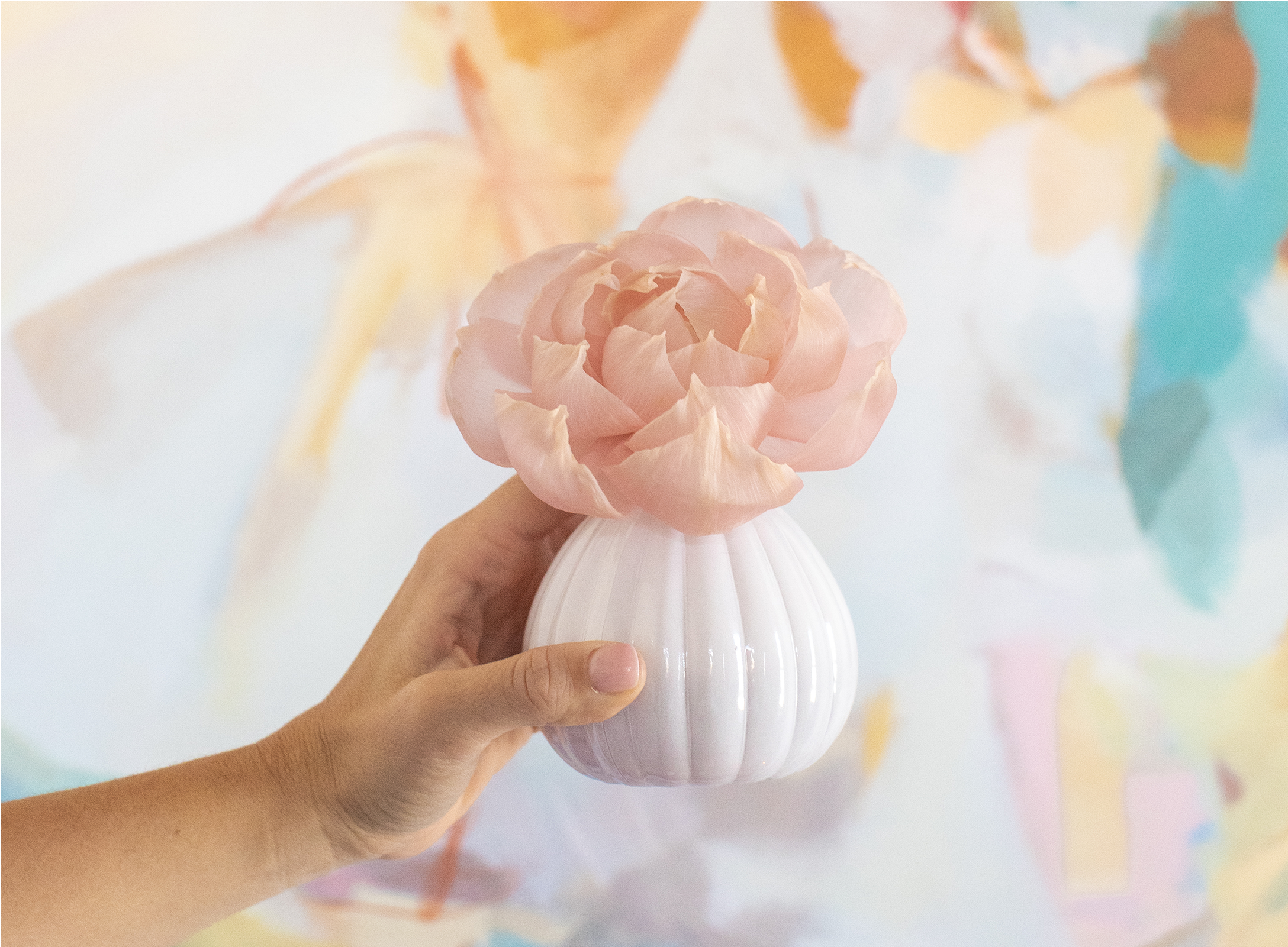 A hand holds a small white vase with a single large, pale pink flower in front of a colorful, abstract pastel background.