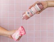 A hand pours pink Sweet Grace liquid onto a pink and white foamy sponge, held by another hand, against a pink tiled background.