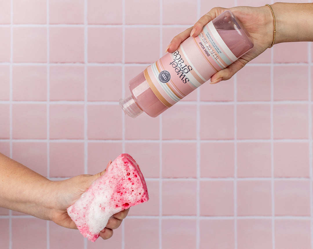 A hand pours pink Sweet Grace liquid onto a pink and white foamy sponge, held by another hand, against a pink tiled background.