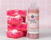 Three pink sponges stacked with soap suds beside a bottle of Sweet Grace dish soap on a counter, against a pink tiled background.
