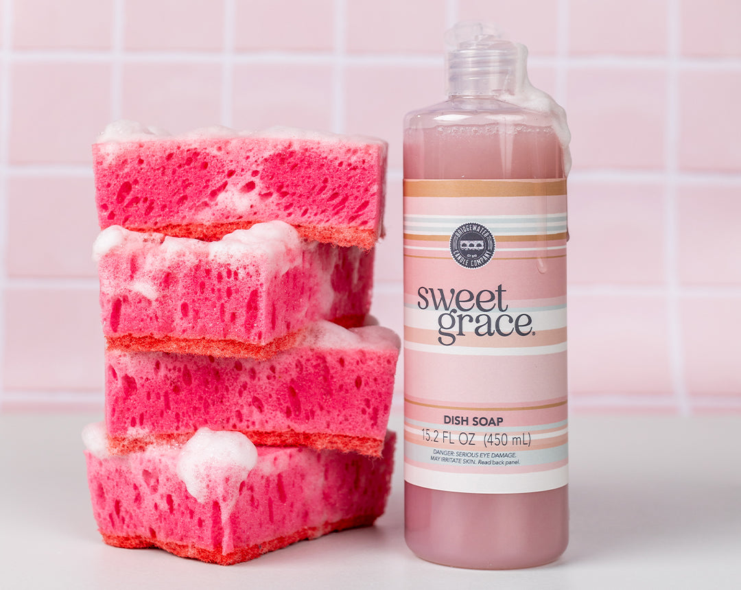 Three pink sponges stacked with soap suds beside a bottle of Sweet Grace dish soap on a counter, against a pink tiled background.