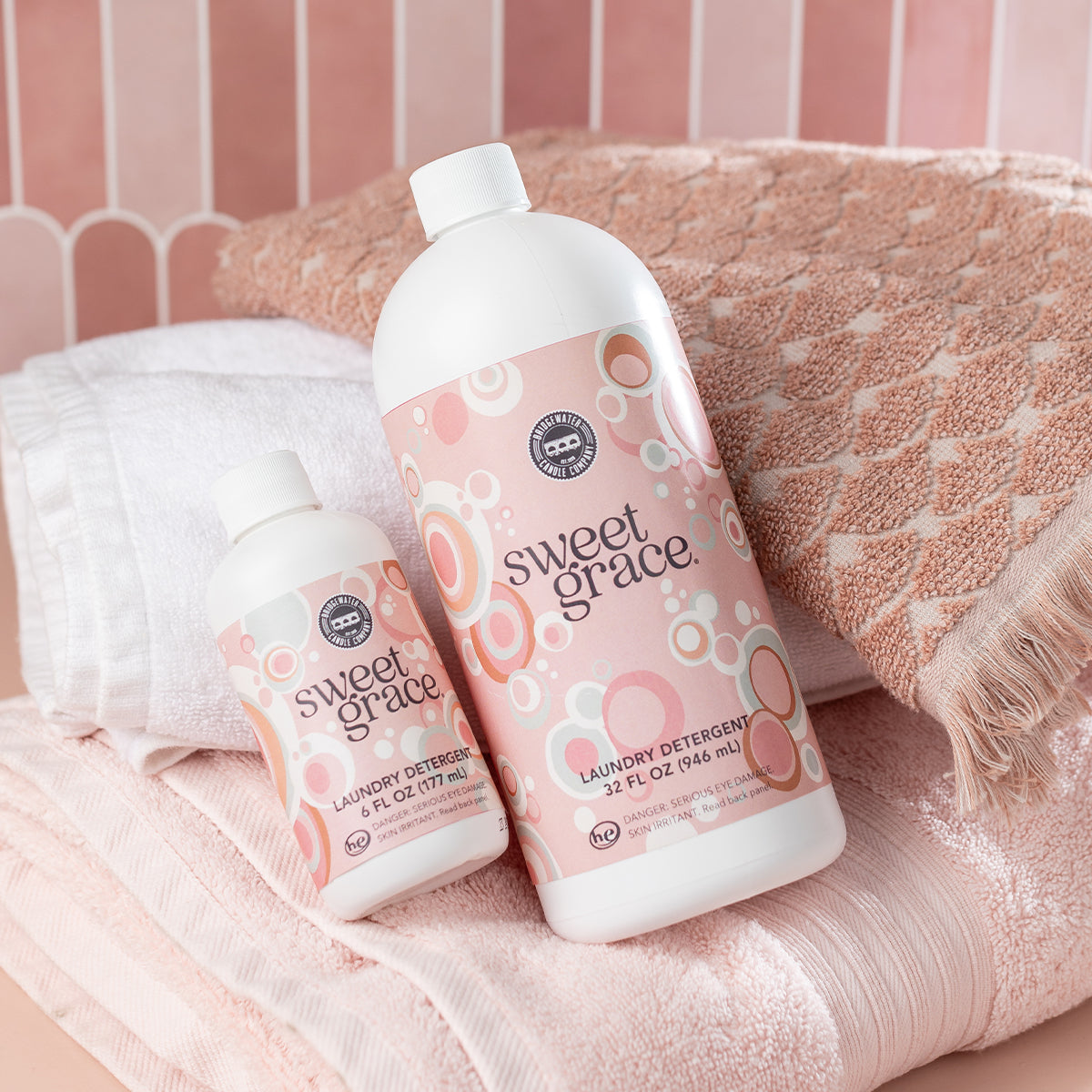 Two bottles of Sweet Grace laundry detergent, one large and one small, are placed on folded white and peach towels against a pink tiled background. The detergent bottles have a pink, white, and peach bubble design.