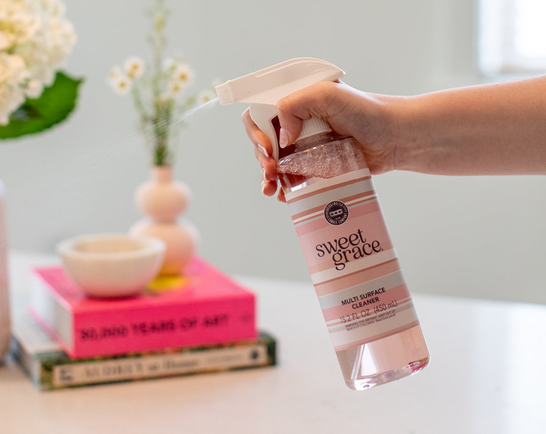 A hand sprays a bottle of Sweet Grace multi-surface cleaner in a bright room with pink books, a bowl, and a vase of flowers on a table in the background.