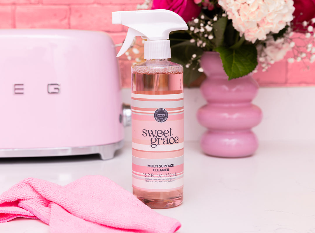 A bottle of Sweet Grace multi-surface cleaner sits on a white countertop with a pink cloth, pink toaster, and pink vase with flowers in the background.