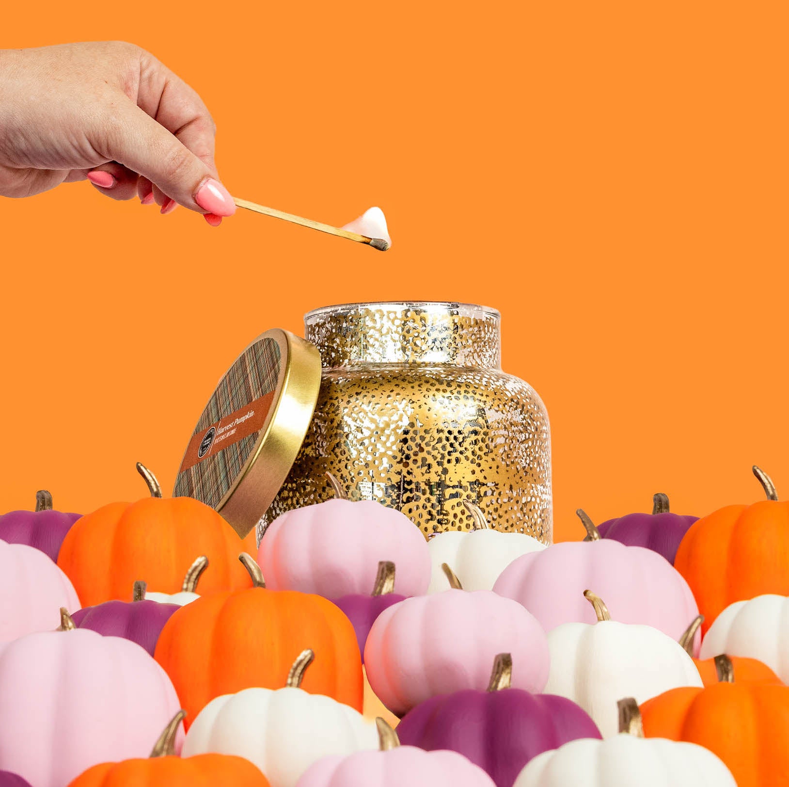 A hand lights a match above the Bridgewater Candle Company Harvest Pumpkin Mercury Jar Candle, a gold-speckled soy-blend wax candle jar surrounded by mini pumpkins in orange, white, pink, and purple against an orange background.