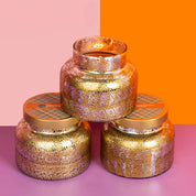 Three Bridgewater Candle Company Harvest Pumpkin Mercury Jar Candles—each featuring gold and pink speckled glass filled with luxurious soy-blend wax—are stacked in a pyramid, with two lids at the base, set against a vibrant pink and orange backdrop. Perfect for evoking cozy autumn evenings, these candles infuse any space with warm harvest pumpkin notes.
