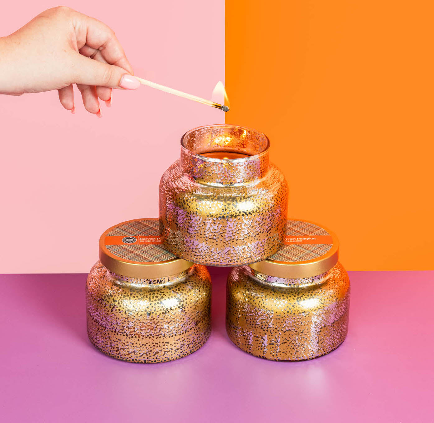 A hand lights the Bridgewater Candle Company Harvest Pumpkin Mercury Jar Candle with a match. Made from soy-blend wax, the gold-speckled candle sits atop two identical jars on a purple surface, set against a pink and orange background.