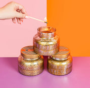 A hand lights the Bridgewater Candle Company Harvest Pumpkin Mercury Jar Candle with a match. Made from soy-blend wax, the gold-speckled candle sits atop two identical jars on a purple surface, set against a pink and orange background.