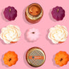 A flat lay of fall decor on a pink background, with purple, orange, and pink pumpkins, white faux flowers, and a lit Bridgewater Candle Company Harvest Pumpkin Holiday Mercury Jar Candle in a copper jar.