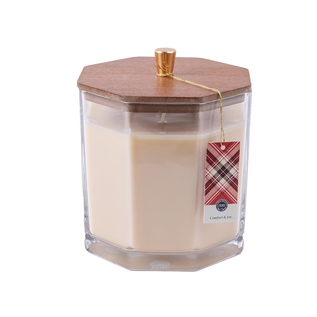 The Bridgewater Candle Company Comfort & Joy Holiday Large Decorative Candle features a cream soy-blend wax in a clear hexagonal glass jar with a wooden lid, gold knob, and red plaid label hanging from a gold string. Premium fragrance. White background.