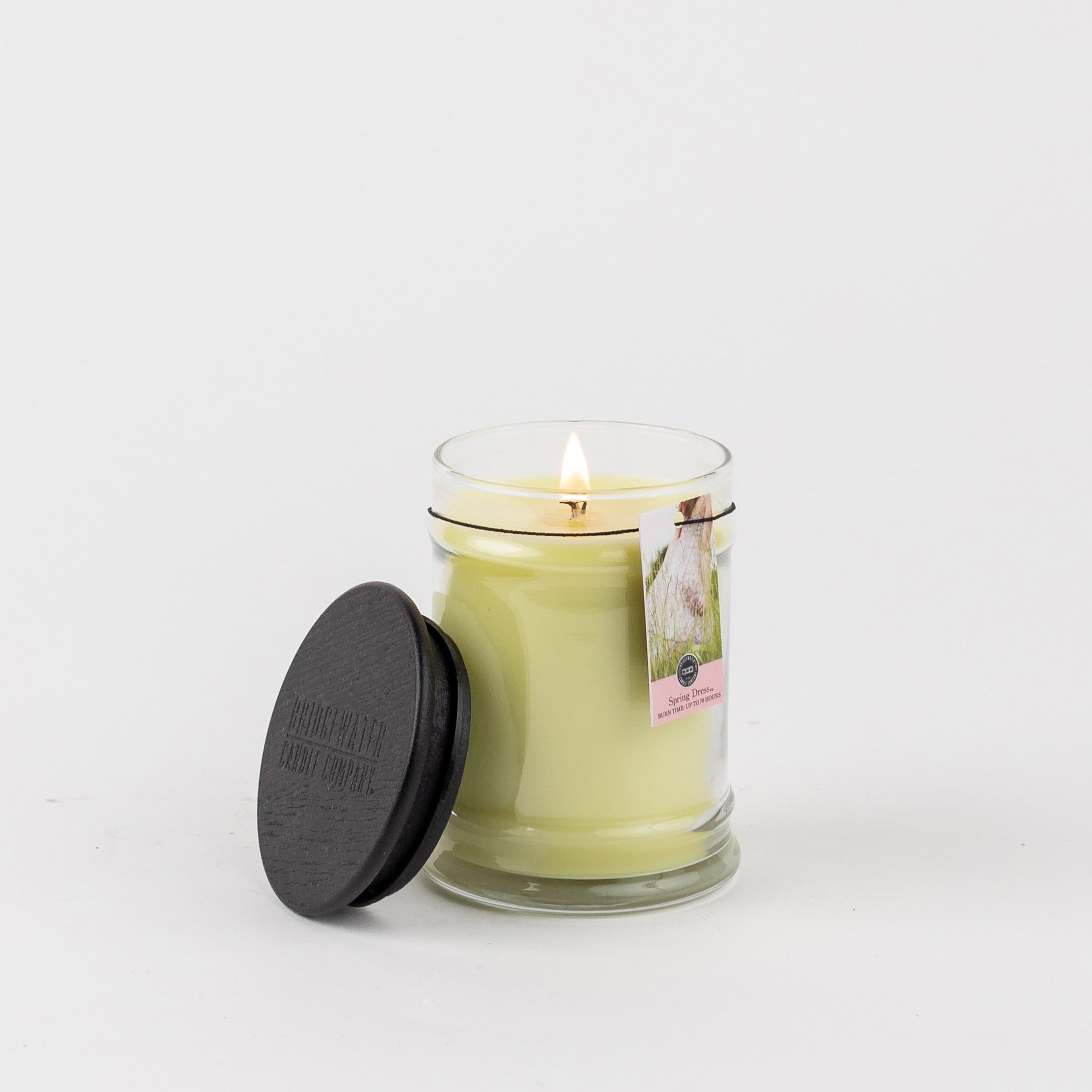 A lit Spring Dress 8oz Jar Candle by Bridgewater Candle Company sits in a clear glass jar with a black lid nearby and a tag attached, all against a plain white background.