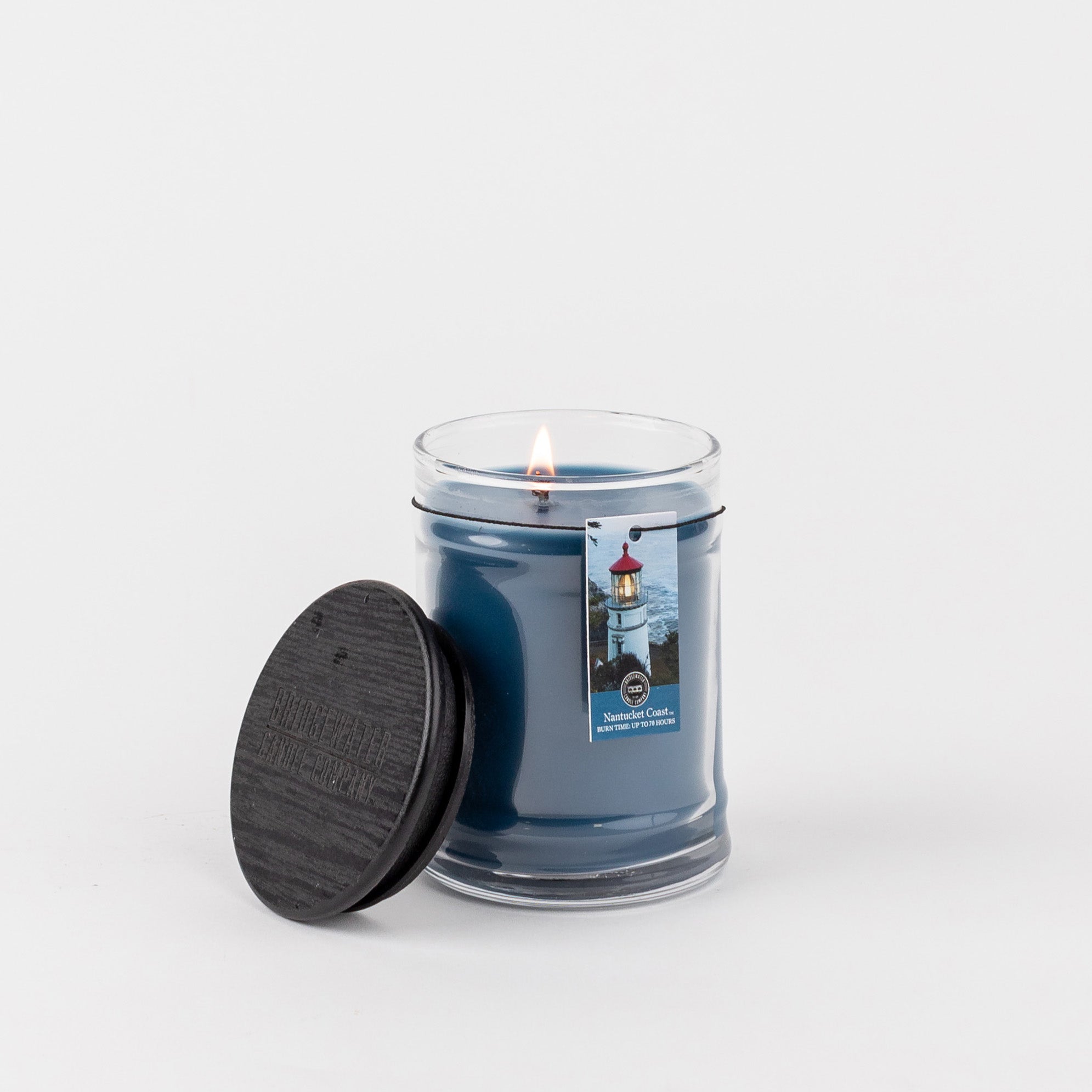 A lit Nantucket Coast 8oz Jar Candle by Bridgewater Candle Company in clear glass with a black wooden lid beside it. The label shows a lighthouse, reflecting the fresh coastal scent, all set against a plain white background.