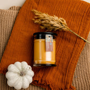The Autumn Gold Small Jar Candle by Bridgewater Candle, featuring an orange label, sits on an orange cloth next to a white decorative pumpkin and dried wheat on brown textured fabric. Made with soy-blend wax and filled with signature Bridgewater fragrance.