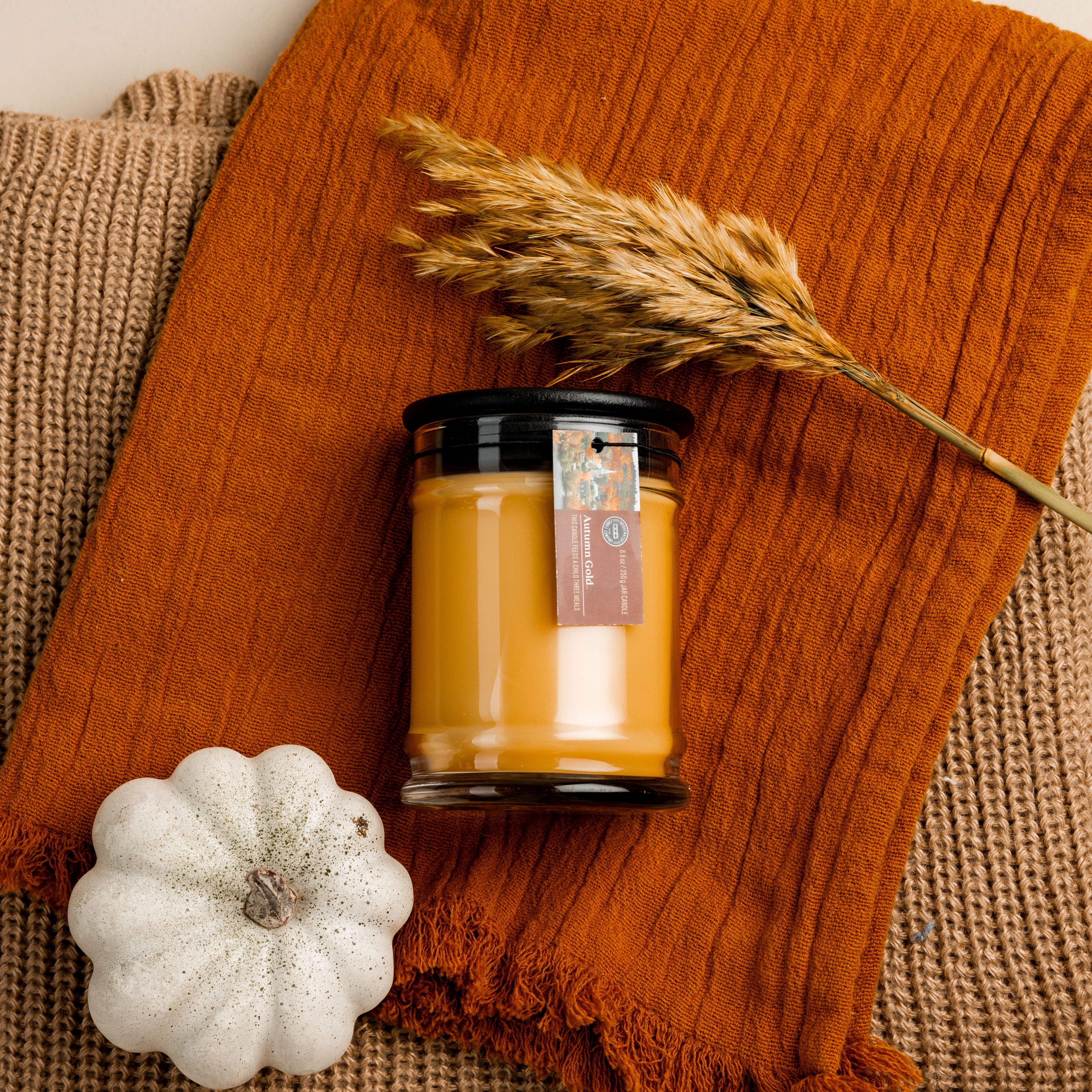 The Autumn Gold Small Jar Candle by Bridgewater Candle, featuring an orange label, sits on an orange cloth next to a white decorative pumpkin and dried wheat on brown textured fabric. Made with soy-blend wax and filled with signature Bridgewater fragrance.