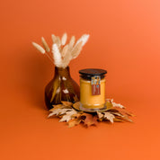 A glass vase with dried grass stems and a Bridgewater Candle Autumn Gold Small Jar Candle on a gold plate, surrounded by artificial autumn leaves, set against an orange background.
