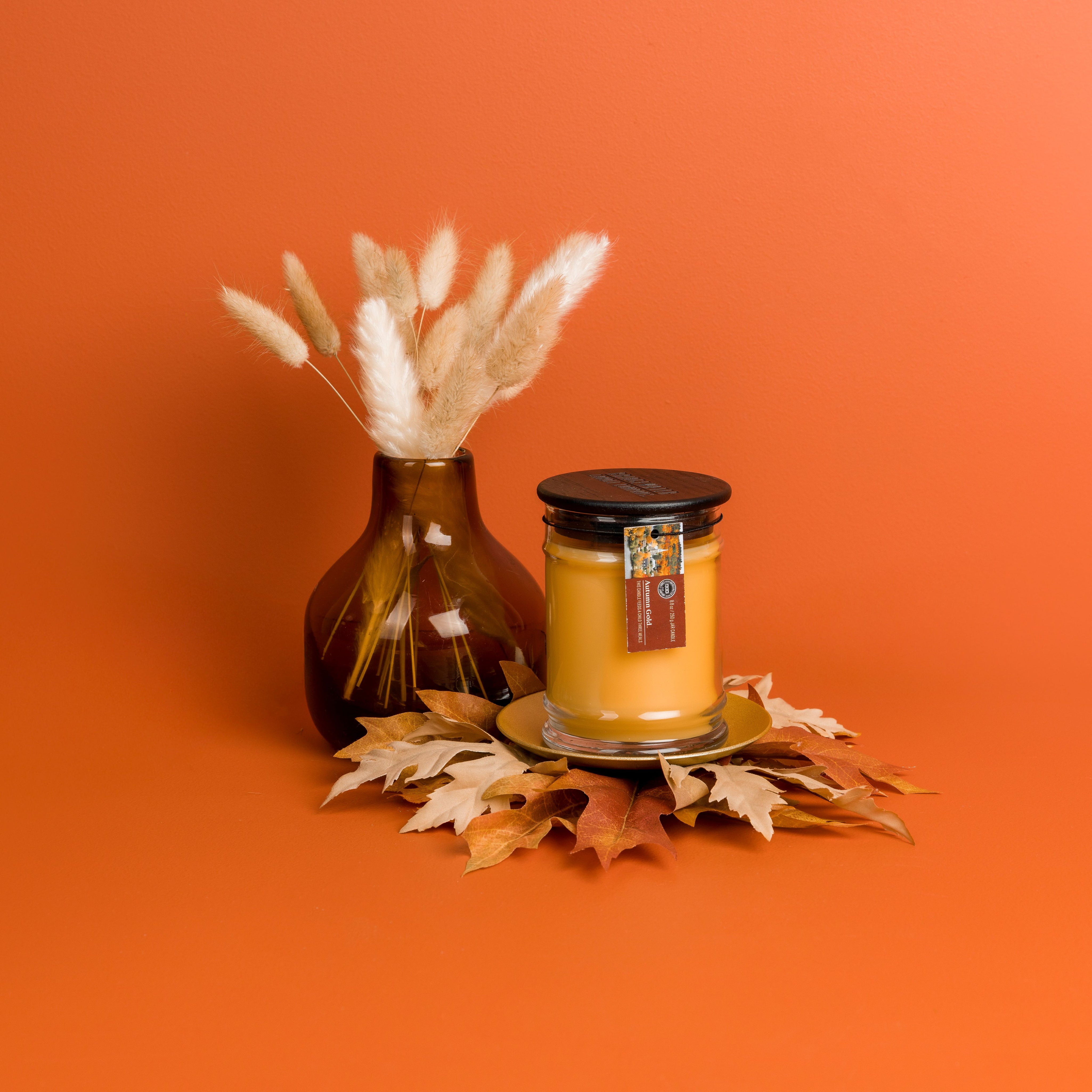 A glass vase with dried grass stems and a Bridgewater Candle Autumn Gold Small Jar Candle on a gold plate, surrounded by artificial autumn leaves, set against an orange background.