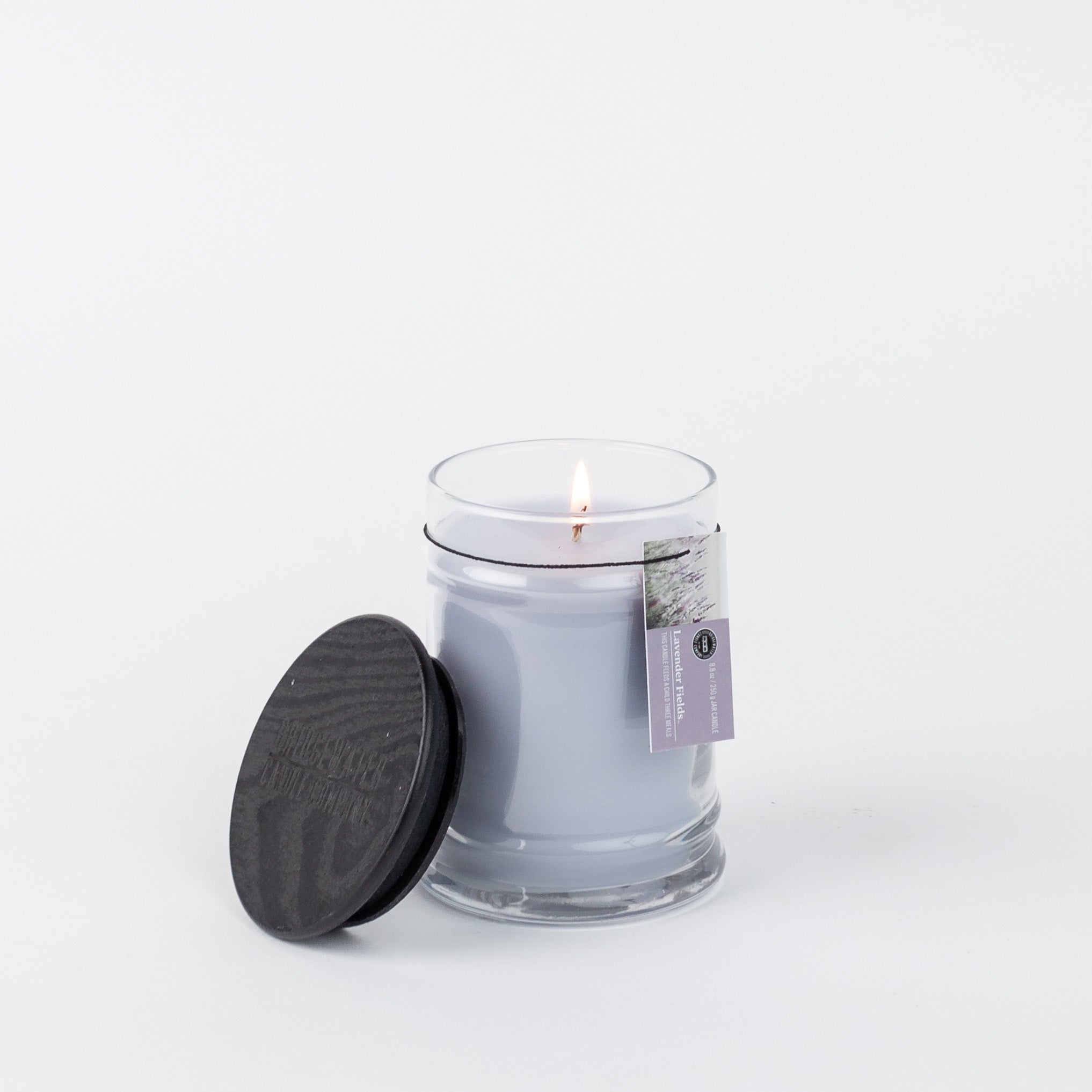A lit Bridgewater Candle Company Lavender Fields 8oz Small Jar Candle sits in a clear jar with its black wooden lid nearby on a white background. A small label is attached to the jar.