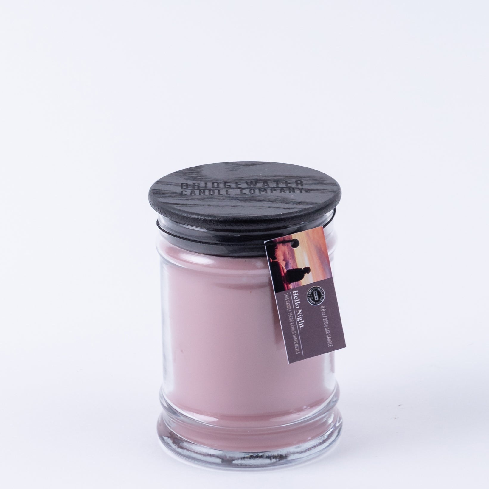 The Bridgewater Candle Company Hello Night 8oz Jar Candle features pink soy-blend wax, a black lid, and a sunset tag with two people. Its white background makes it perfect for enjoying signature Bridgewater fragrance.