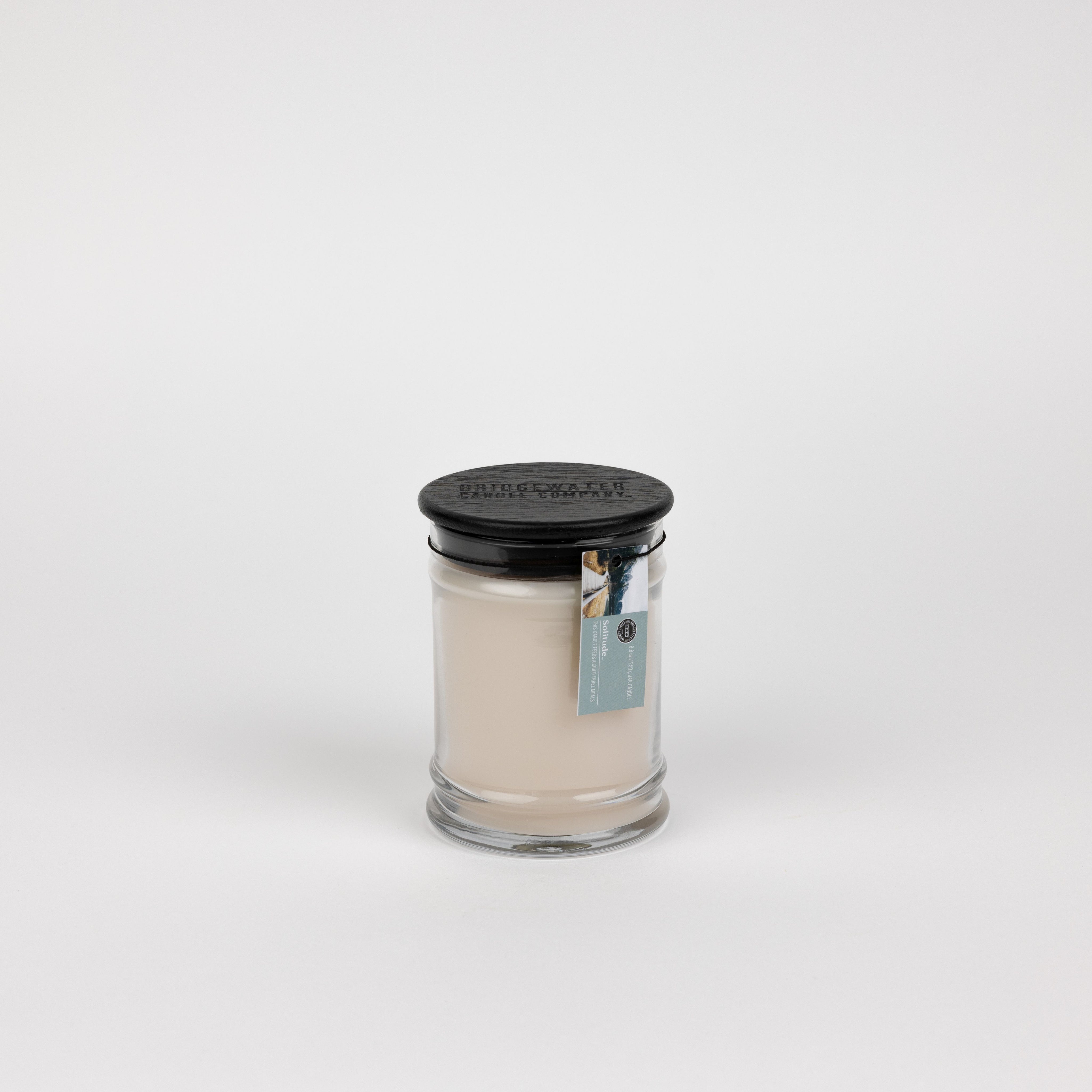 The Solitude Small Jar Candle by Bridgewater Candle features off-white soy-blend wax in a clear glass jar with a black lid and a blue rectangular tag, displayed on a plain white background.