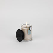 A lit Solitude Small Jar Candle by Bridgewater Candle, filled with soy-blend wax, sits in a clear glass jar with a black lid beside it. A blue tag is attached, set against a plain white background to highlight its elegant fragrance.