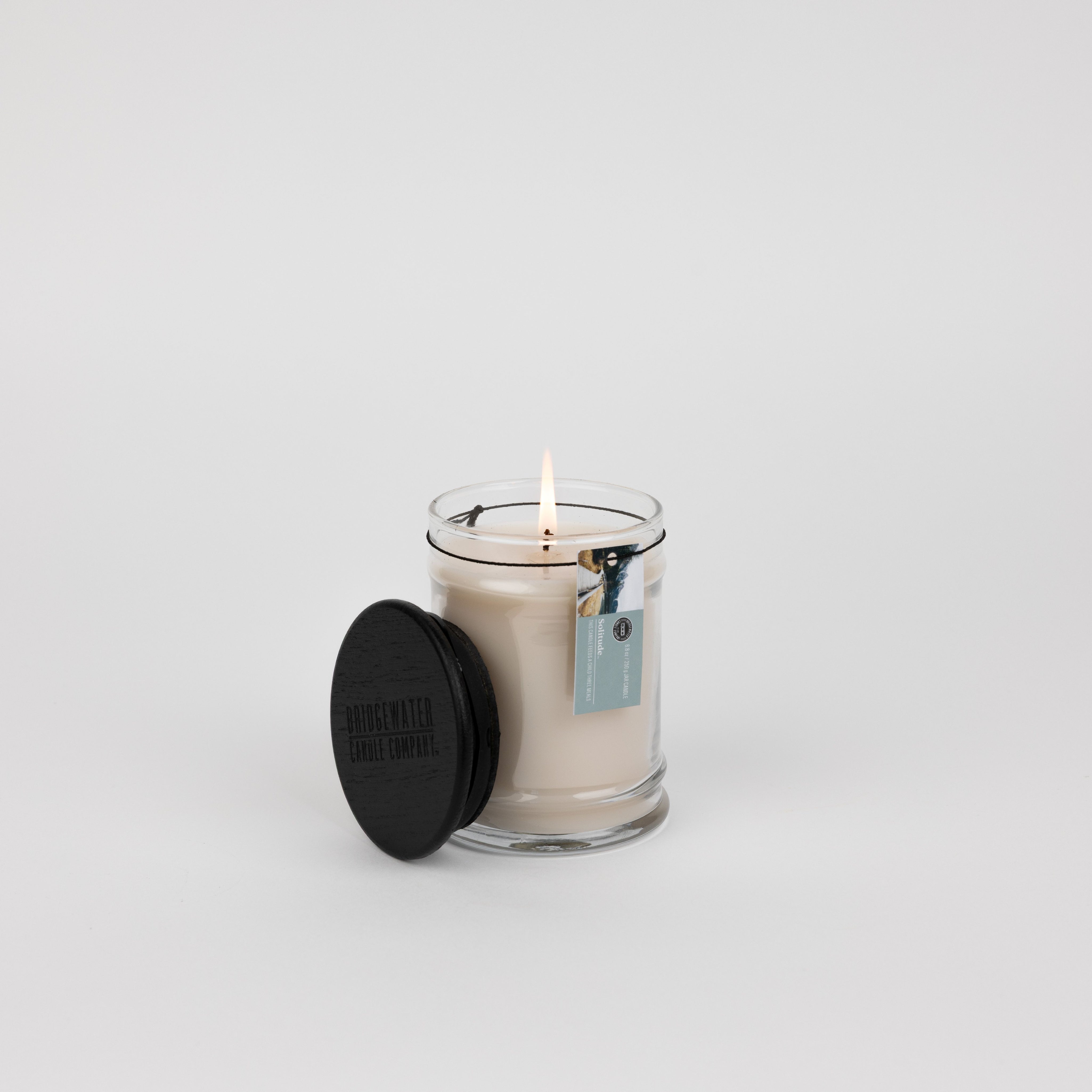 A lit Solitude Small Jar Candle by Bridgewater Candle, filled with soy-blend wax, sits in a clear glass jar with a black lid beside it. A blue tag is attached, set against a plain white background to highlight its elegant fragrance.