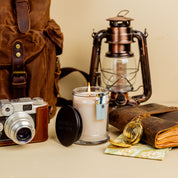 A still life of travel items includes a vintage camera, a brown backpack, a lit Solitude Small Jar Candle by Bridgewater Candle made with soy-blend wax, a lantern, leather-bound journal, map, and brass compass on a beige surface.